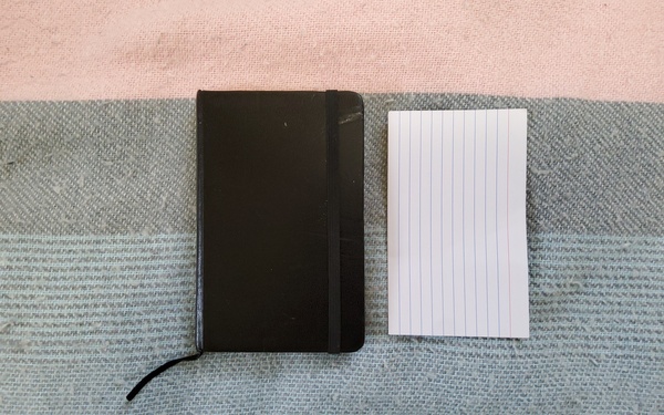 [ID: image of a black, miniature hardcover notebook, next to a 3x5 inch index card. The notebook is barely larger than the card. End ID.]